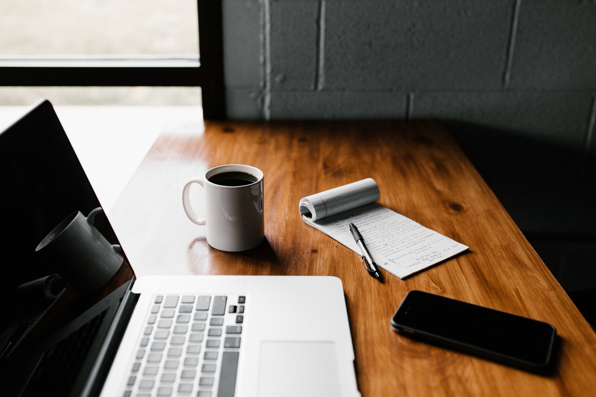 A plain wooden desk with a MacBook, cup of coffee, notebook, and iPhone
