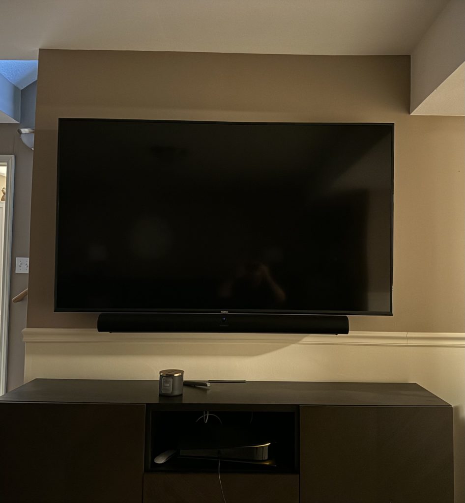 A black TV and soundbar mounted on the wall. Underneath it is a dark brown IKEA Besta storage unit.